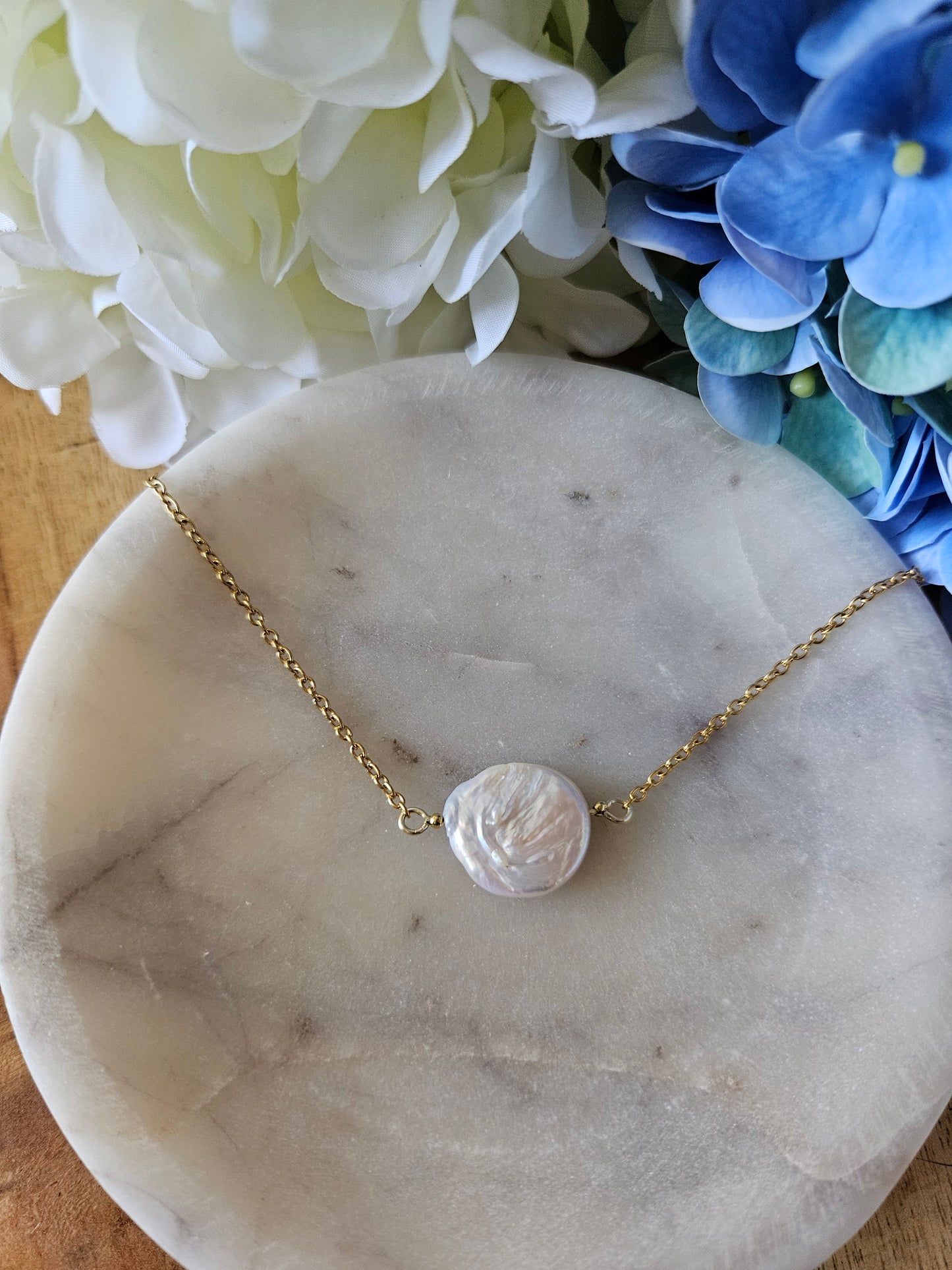 Collar dorado con una delicada cadena de eslabones. El dije es una perla de agua dulce blanca, redonda y de forma orgánica, delicadamente engarzada. El collar descansa sobre una superficie clara.