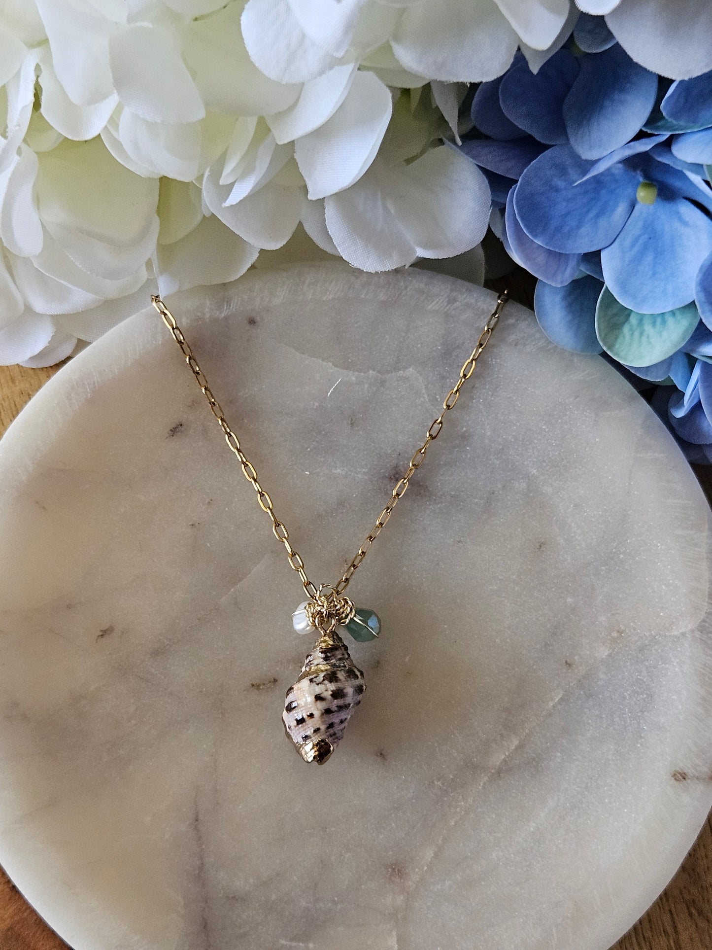 Collar dorado con una cadena de eslabones pequeños. El dije es una concha natural en tonos tierra con detalles dorados, adornada con una perla blanca y un pequeño cristal aguamarina. El collar descansa sobre una superficie de mármol blanca con vetas grises.