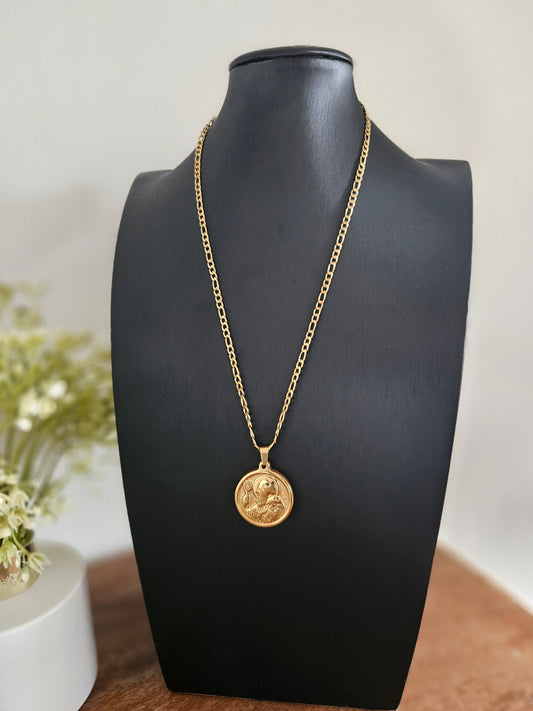 Collar dorado con una cadena de eslabones tipo Figaro. El dije es una medalla redonda dorada con detalles grabados, presumiblemente la medalla de San Benito, colgando de la cadena sobre un busto de maniquí negro.