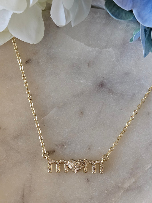 Collar dorado con una fina cadena de eslabones. El dije deletrea la palabra "MOM" con brillantes cristales blancos en micro pavé, con la letra "O" reemplazada por un corazón también brillante.