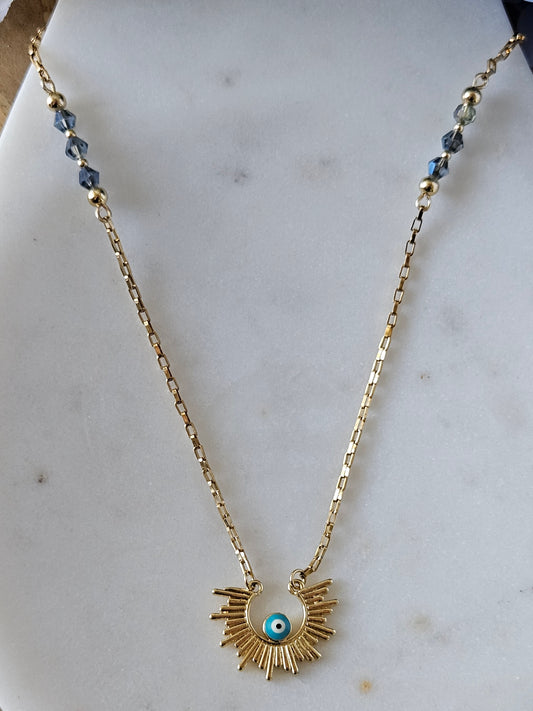 Collar Aura Protectora Mereki: detalle de la cadena "paper clip" dorada con cristales azul ahumado y el dije de ojo turco turquesa con halo dorado.