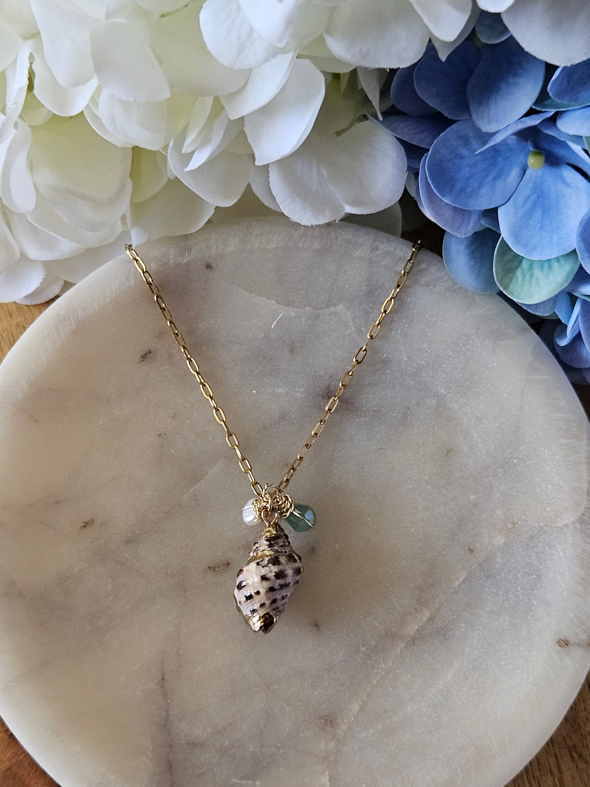Collar dorado con una cadena de eslabones pequeños. El dije es una concha natural en tonos tierra con detalles dorados, adornada con una perla blanca y un pequeño cristal aguamarina. El collar descansa sobre una superficie de mármol blanca con vetas grises.